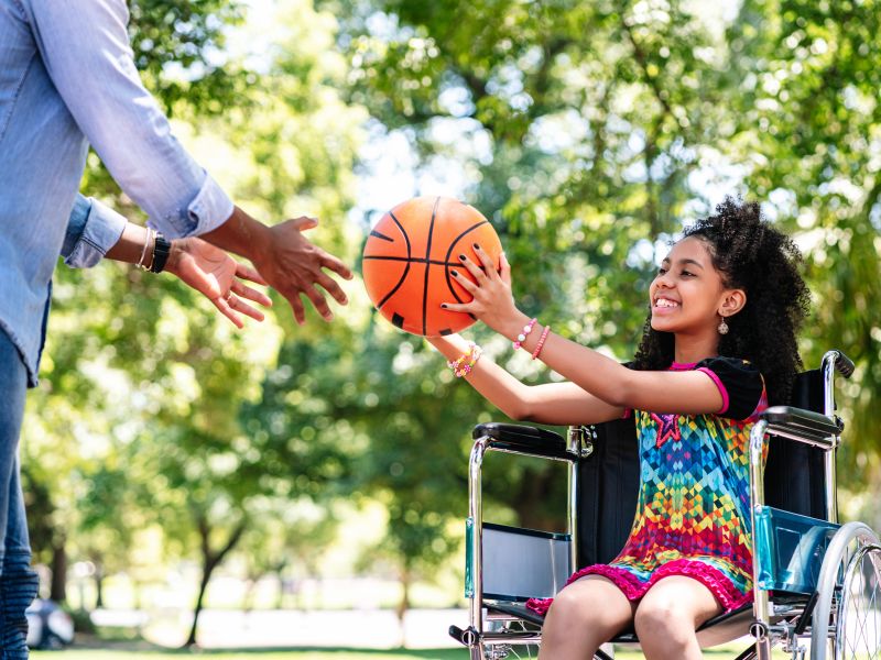 Uma menina em uma cadeira de rodas se divertindo com o pai jogando basquete no parque